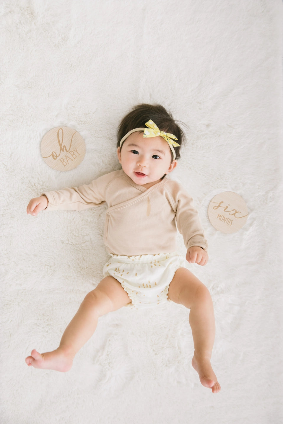 Baby lying on a white surface wearing a beige outfit with a yellow headband, surrounded by circular wooden discs.