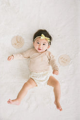 Baby lying on a white surface wearing a beige outfit with a yellow headband, surrounded by circular wooden discs.