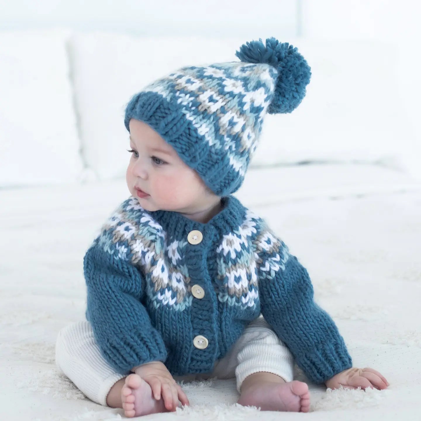 Baby wearing a blue knitted sweater and hat with a pom-pom on a white background