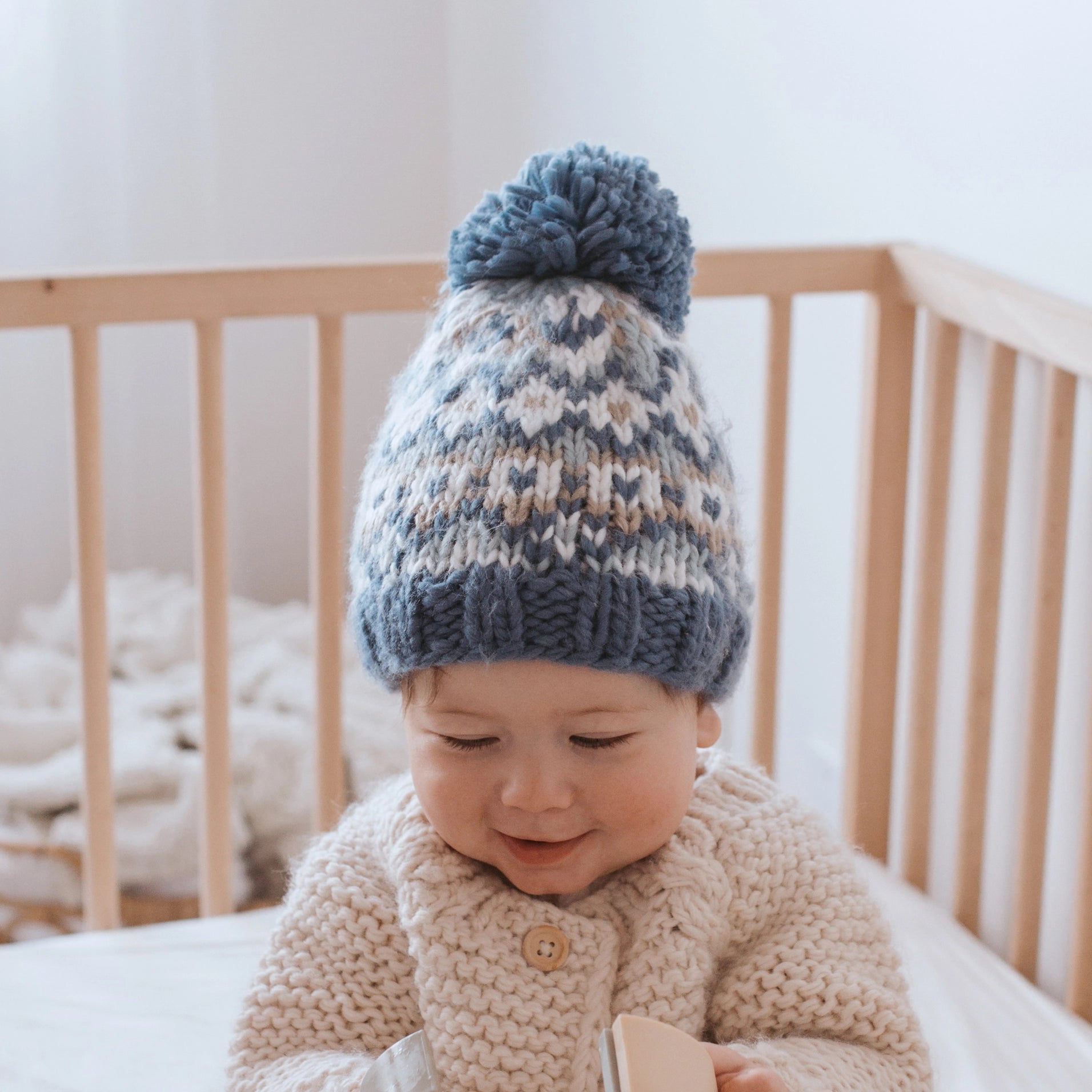 Baby wearing a knitted hat and sweater sitting in a crib.