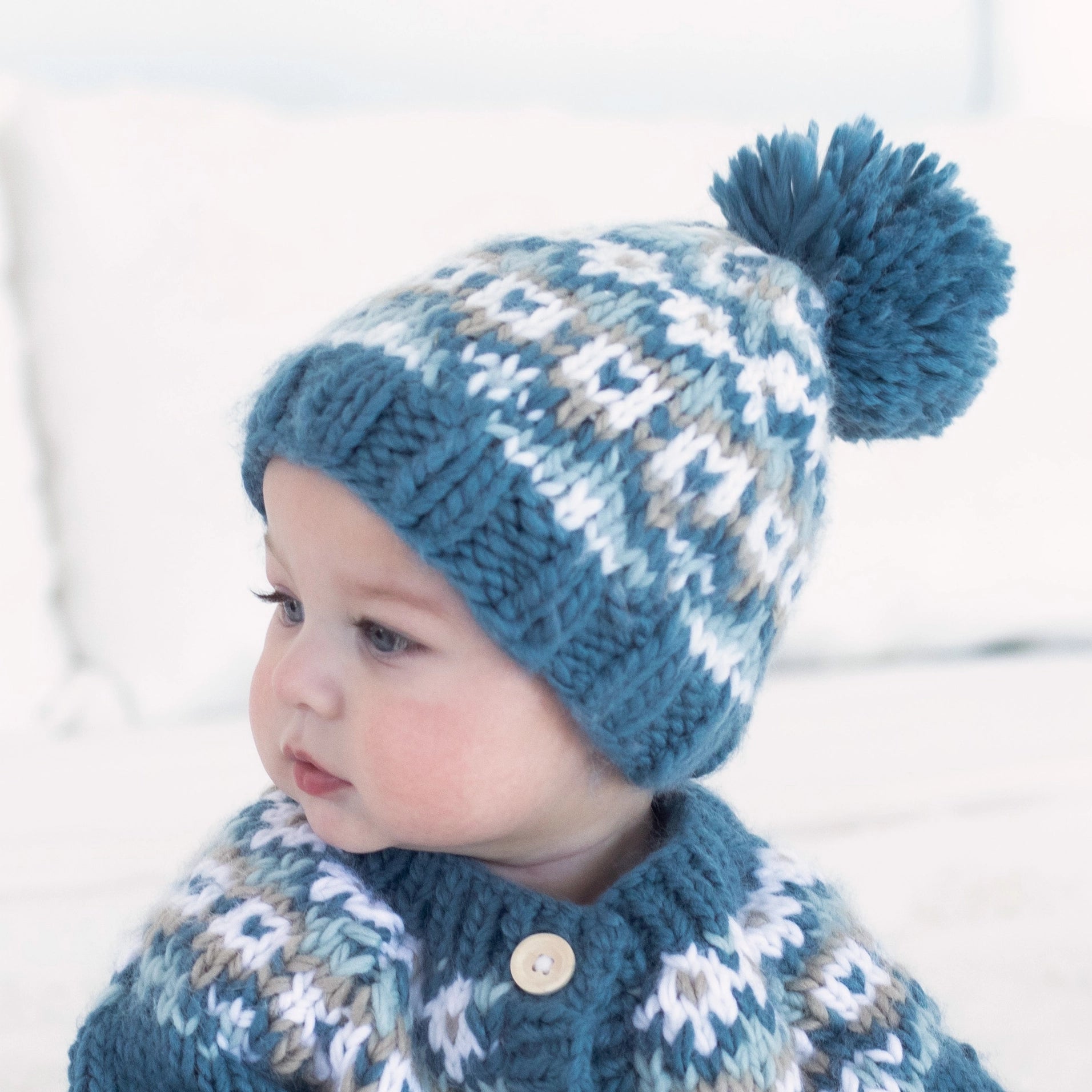 Child wearing a blue knitted hat with a pom-pom and matching cardigan against a light background