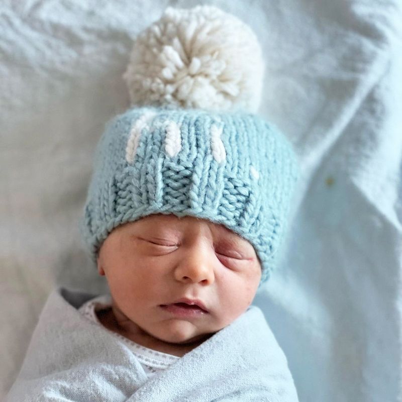 Newborn baby wearing a light blue knitted hat with a white pom-pom on a soft, neutral background.