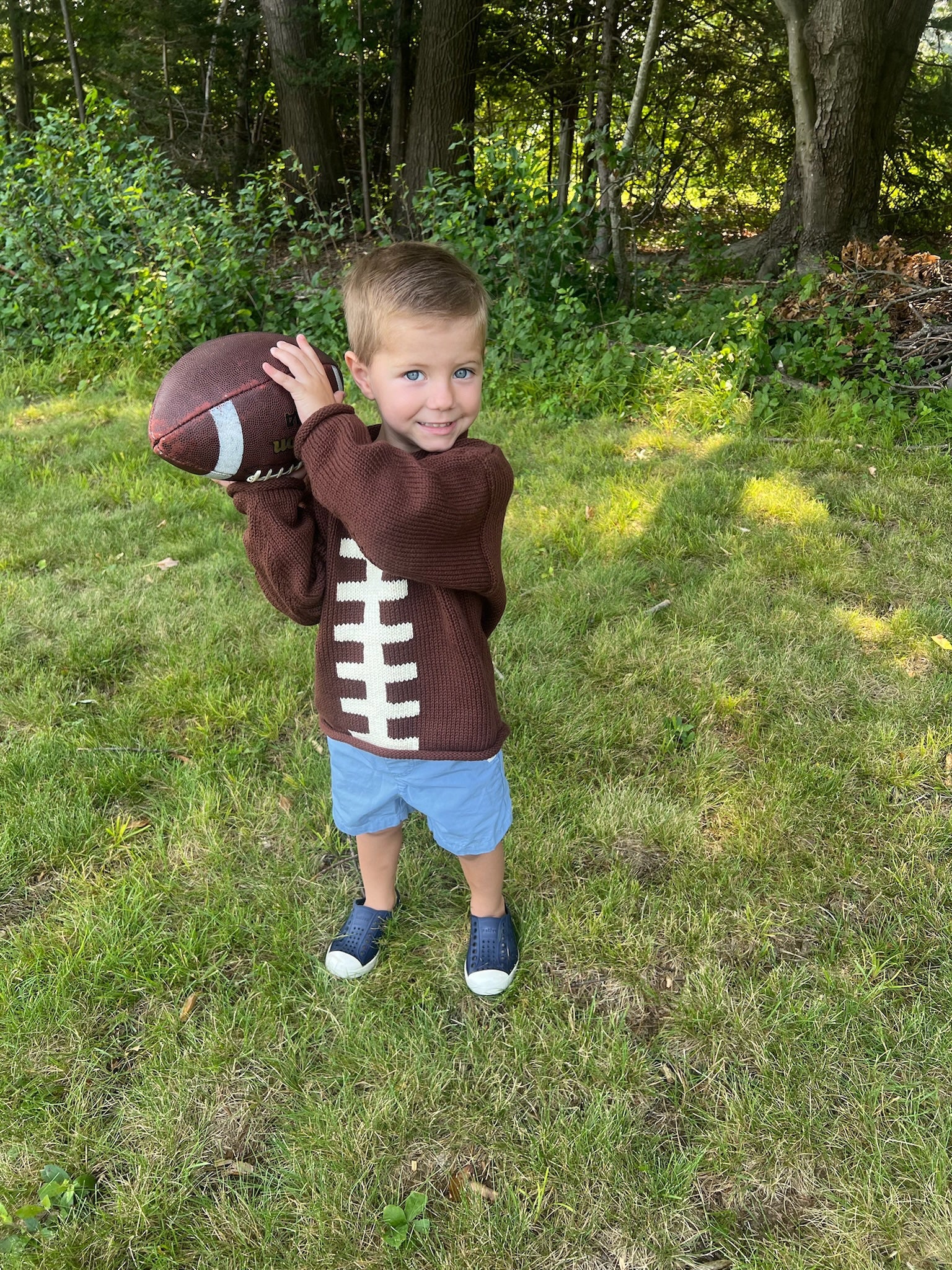 Child wearing a football-themed sweater holding a football outdoors in a grassy area with trees.