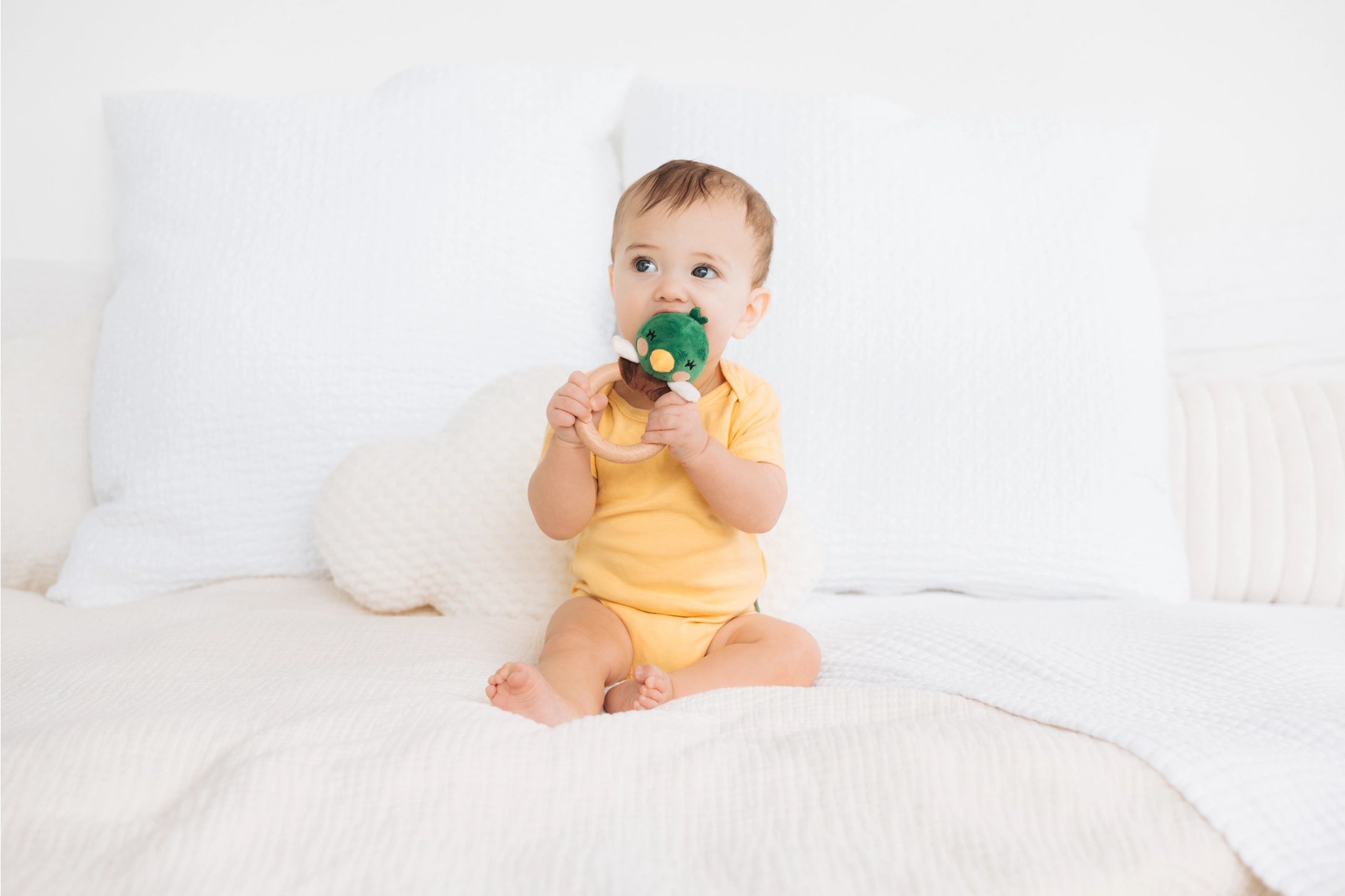 Baby in a yellow outfit holding a green toy on a white couch
