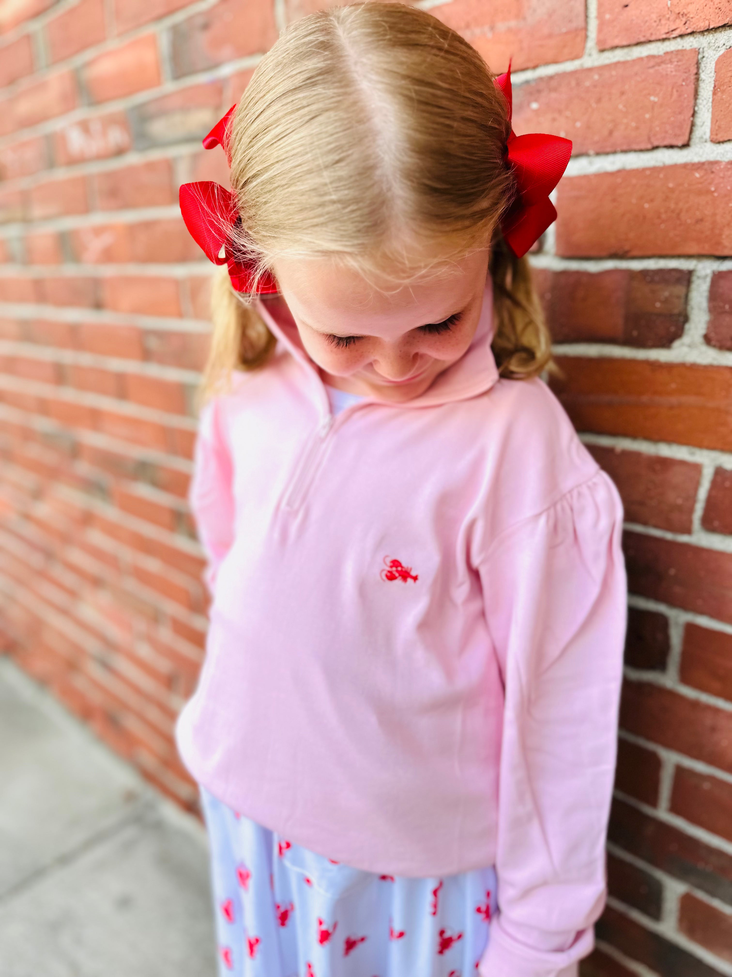 Young girl wearing a pink shirt with a red bow in her hair against a brick wall.