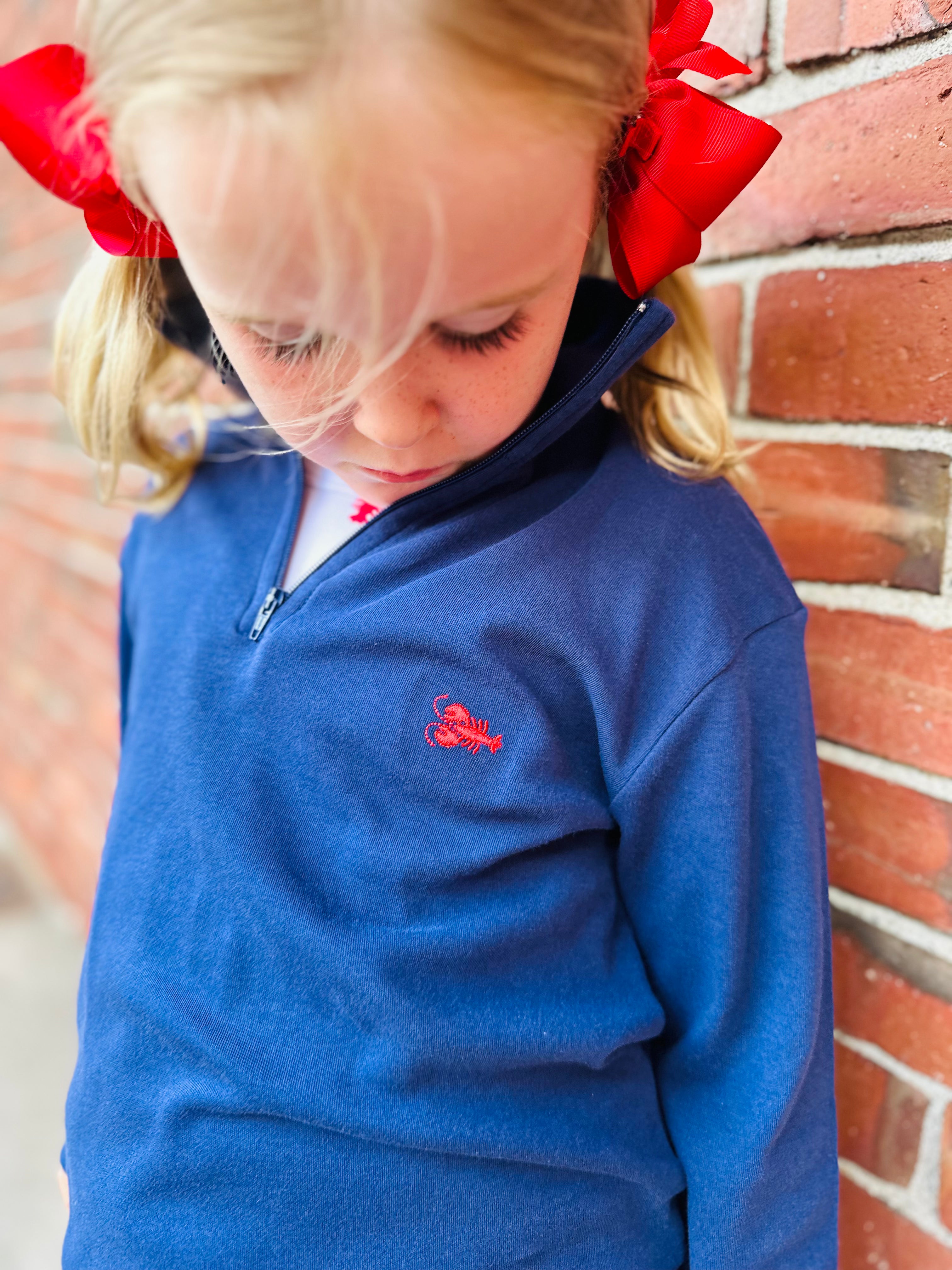 Child wearing a blue sweater with a lobster embroidery, standing against a brick wall.