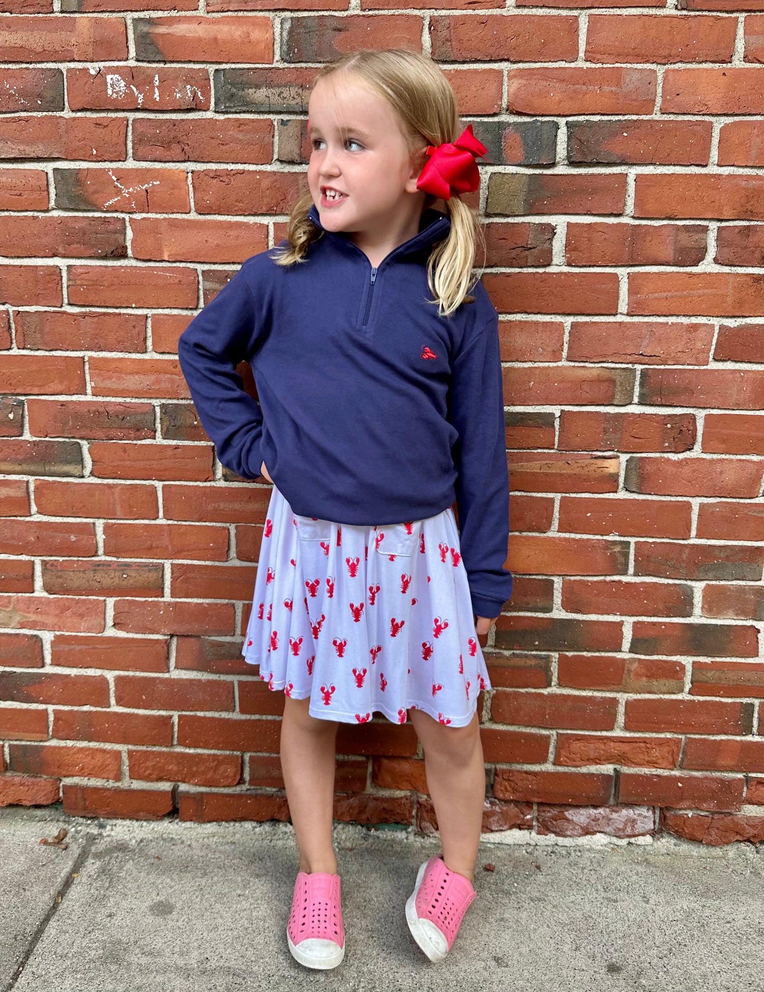 Young girl in a blue top and  lobster patterned skirt standing against a brick wall.