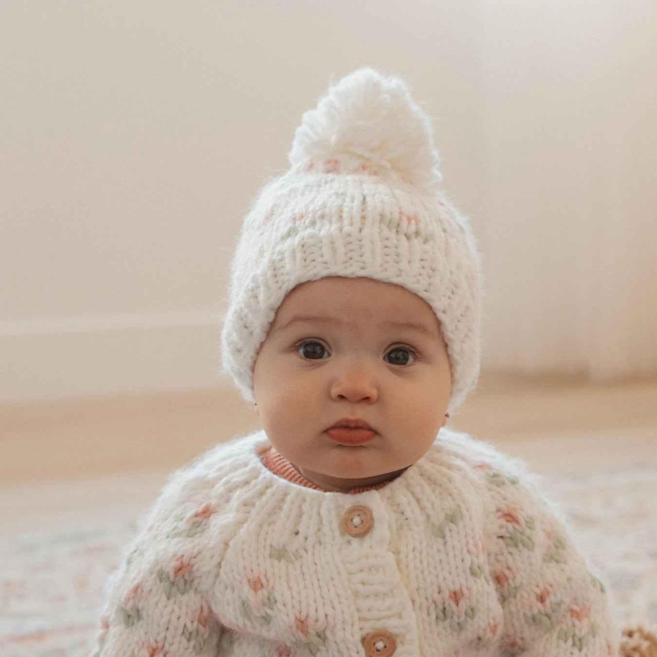 Baby wearing a white knitted outfit with a matching hat on a neutral background