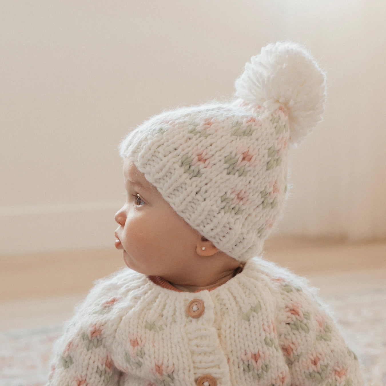 Baby wearing a floral knitted outfit with a white hat featuring a pom-pom on a neutral background