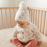 Baby wearing a knitted sweater and hat sitting in a crib.