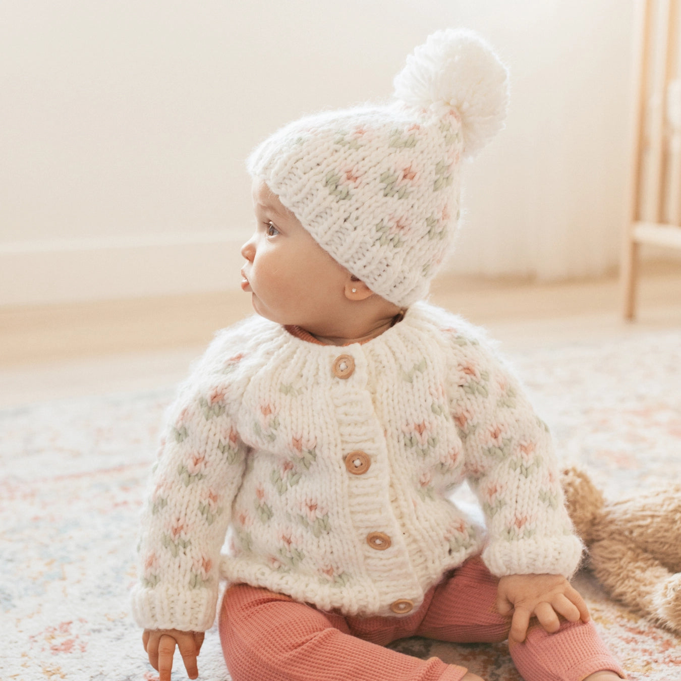 Baby wearing a floral sweater and white knit hat with pom-pom indoors.
