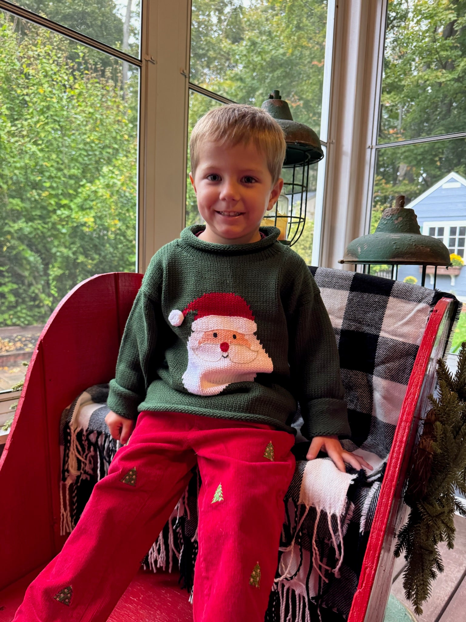 Child wearing a green sweater with a Santa Claus design, sitting on a red chair indoors.