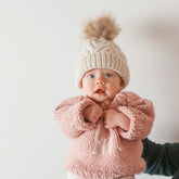 Baby wearing a pink knitted sweater and hat with a pom-pom on a plain background