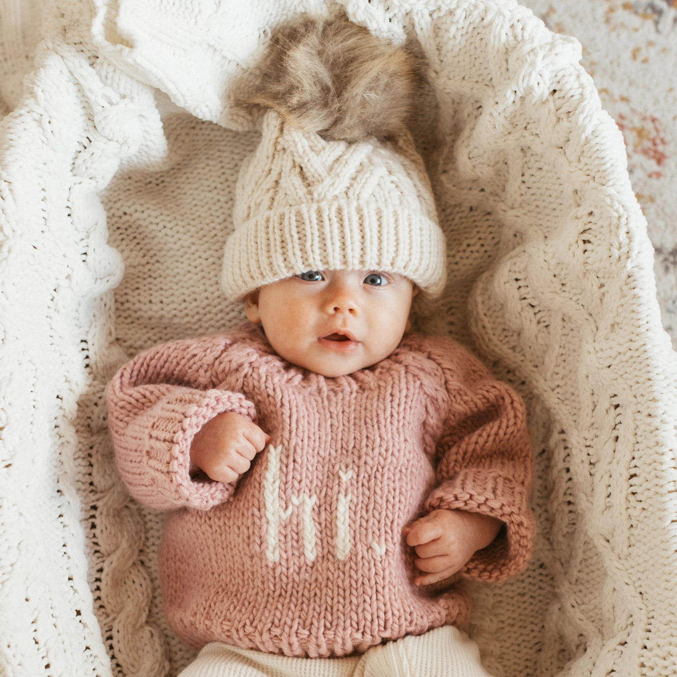 Baby wearing a pink knitted sweater and cream hat lying on a textured blanket.