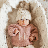 Baby wearing a pink knitted sweater and cream hat lying on a textured blanket.