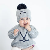 Baby wearing a gray knit hockey sweater and hat with a pom-pom on a white background