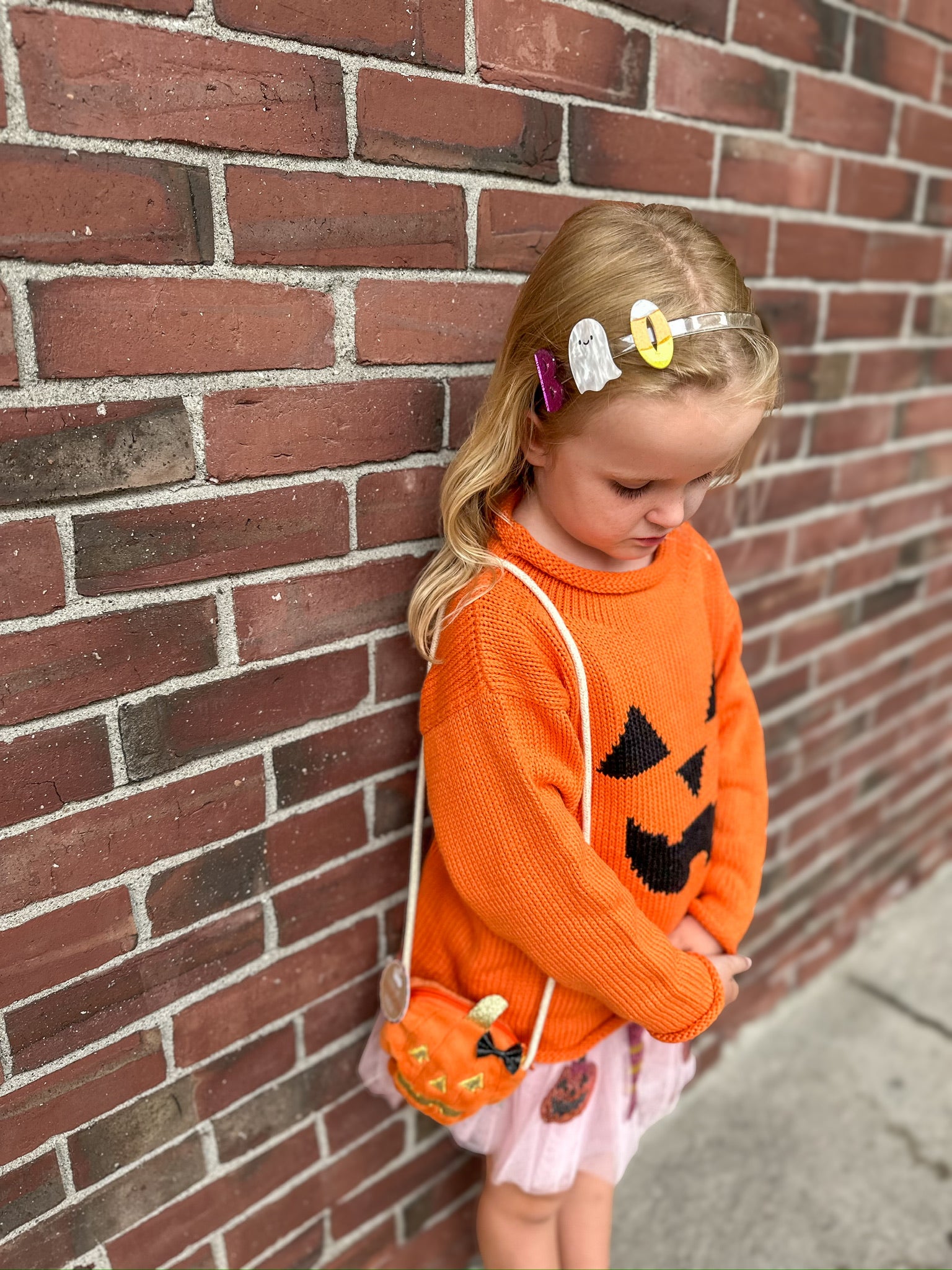 Child wearing an orange sweater with a pumpkin design against a brick wall.
