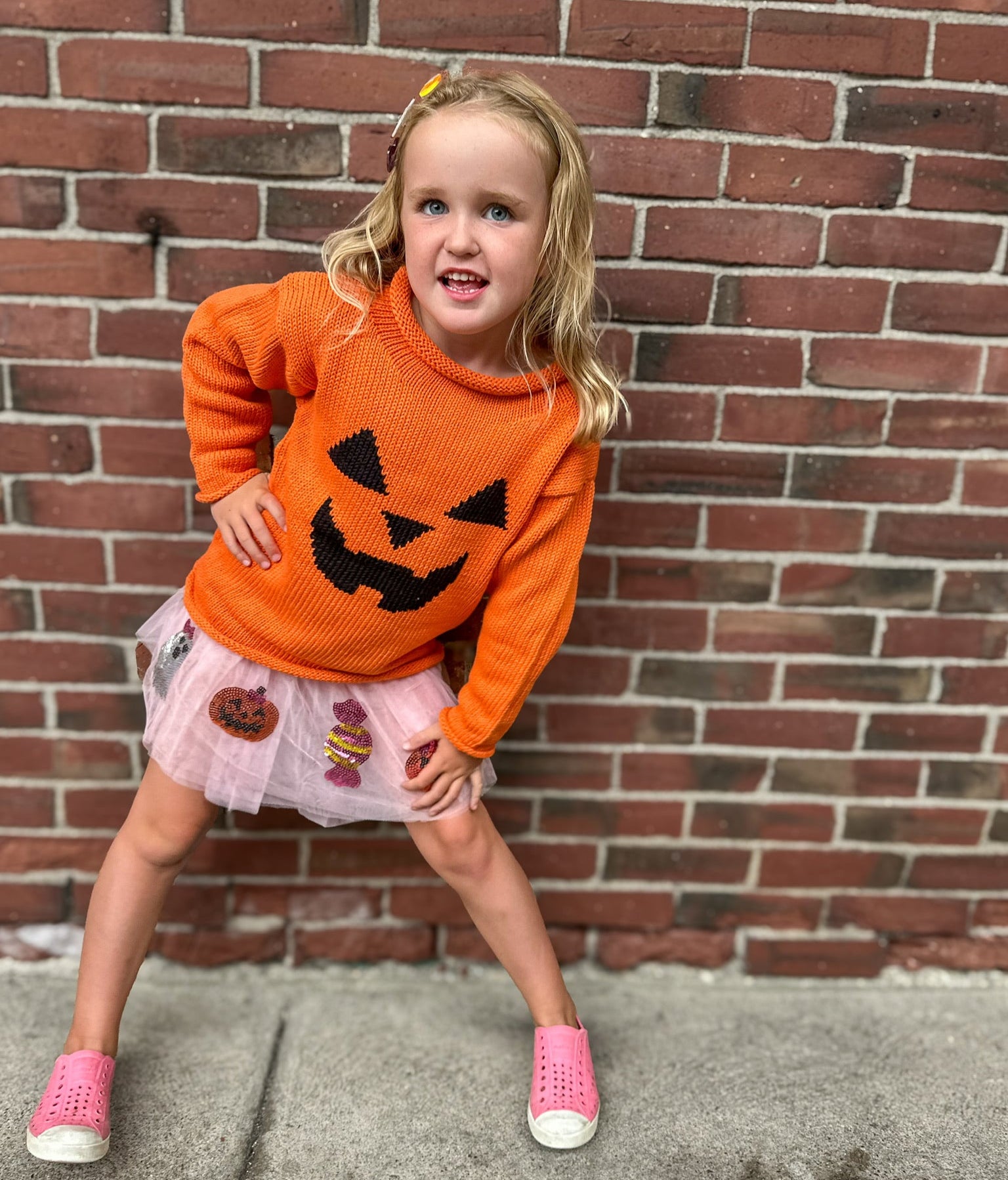 Child wearing an orange sweater with a pumpkin face design, standing against a brick wall.