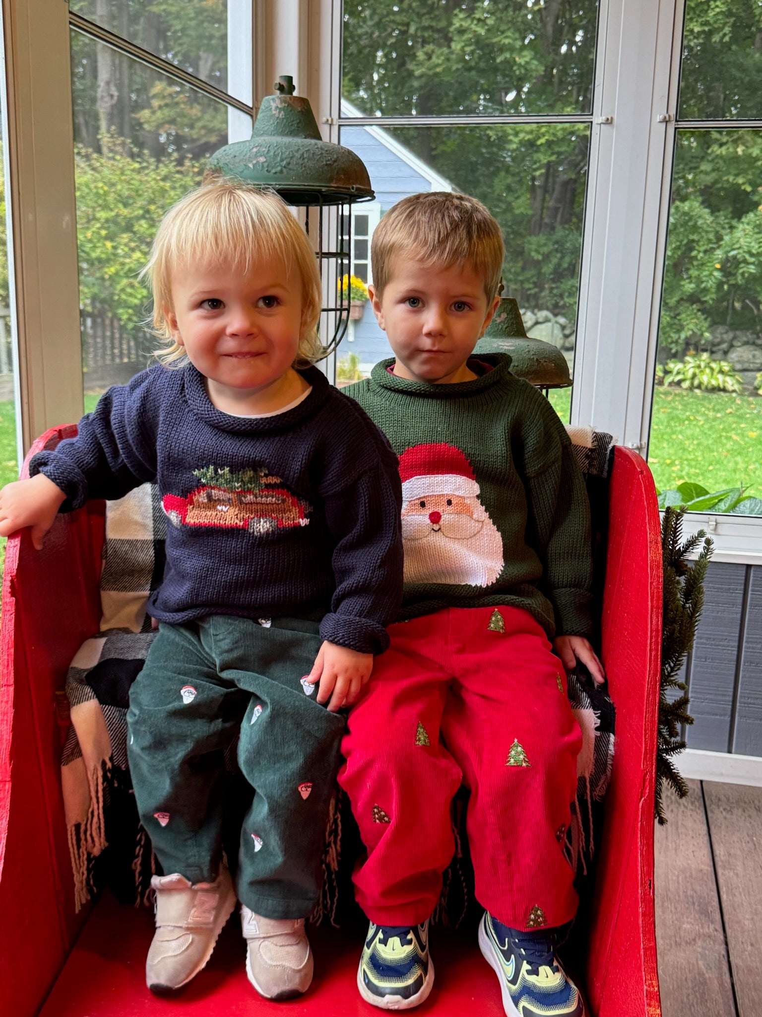 Two children sitting on a red chair wearing Christmas-themed sweaters.