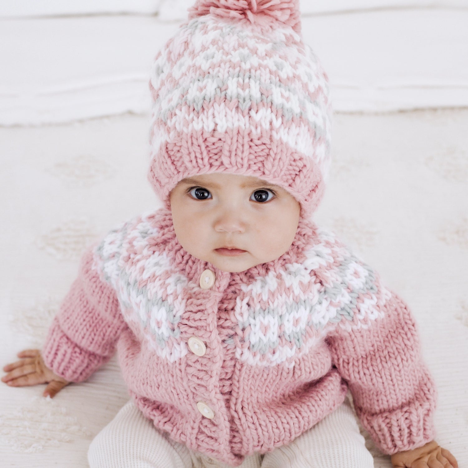 Baby wearing a pink knitted outfit with a matching hat on a light background