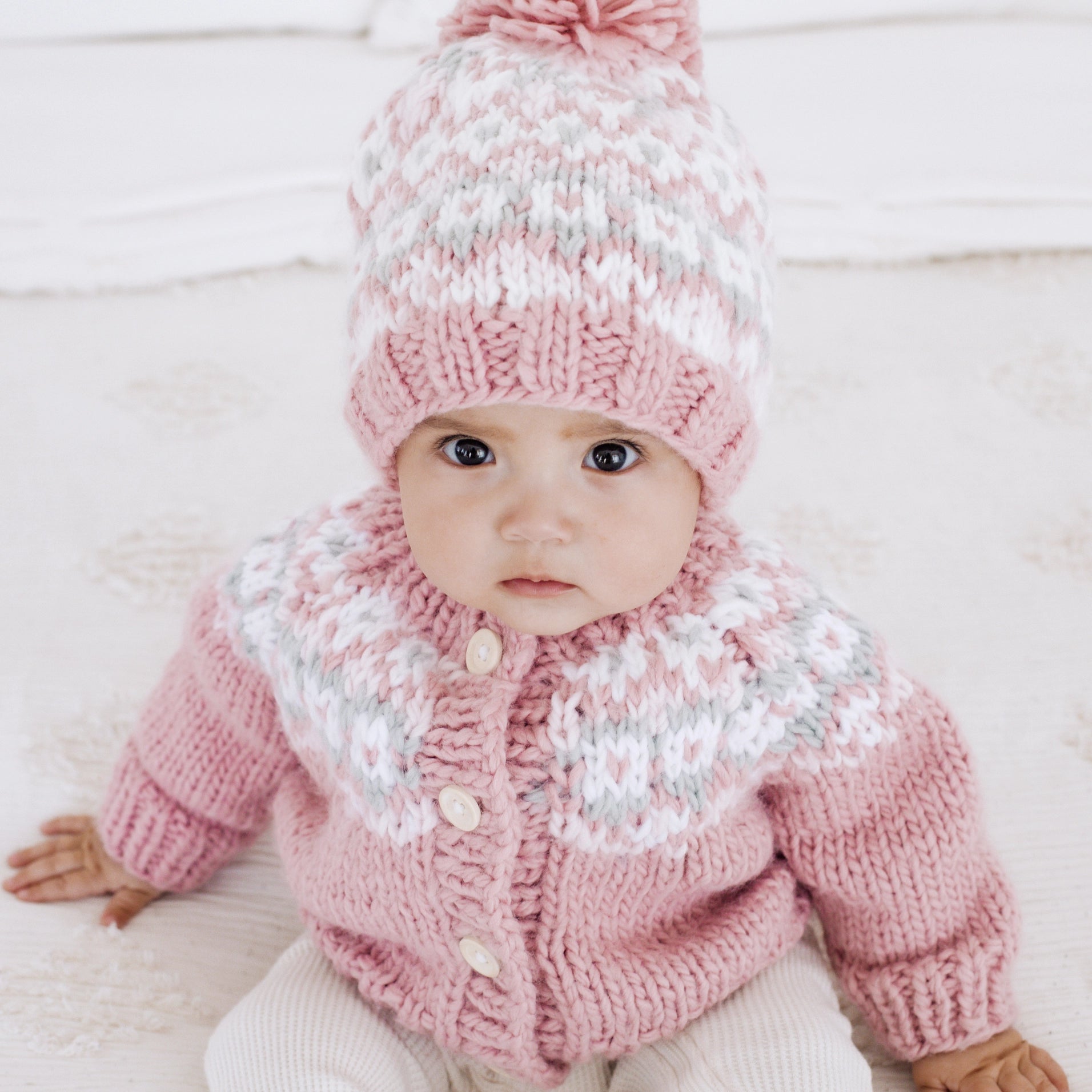 Baby wearing a pink knitted outfit with a matching hat on a light background