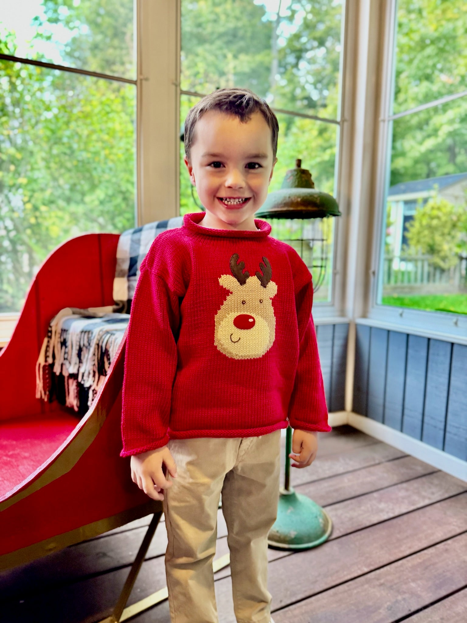 Child wearing a red sweater with a reindeer design on a wooden deck.