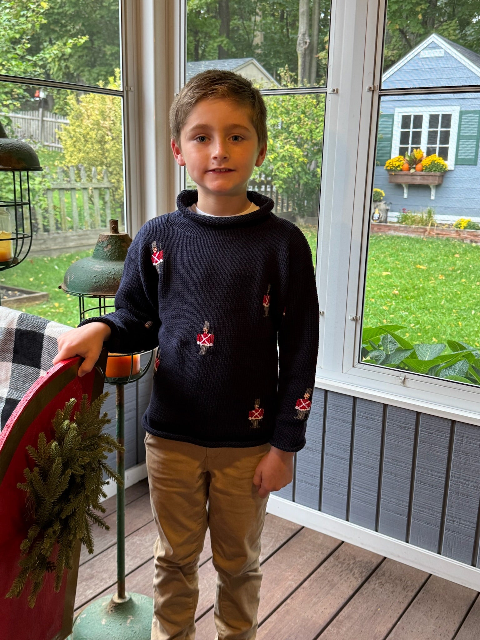 Young boy standing on a wooden deck holding a red object, with a garden and house visible through the window.