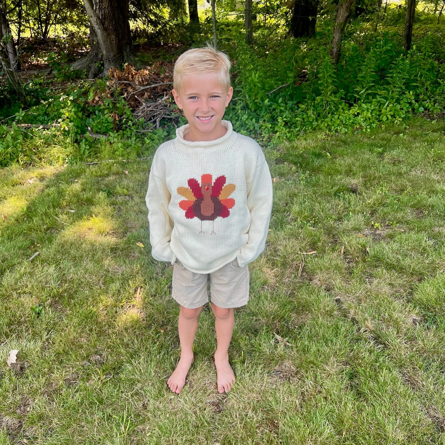 Young boy wearing a sweater with a turkey design outdoors in a grassy area.