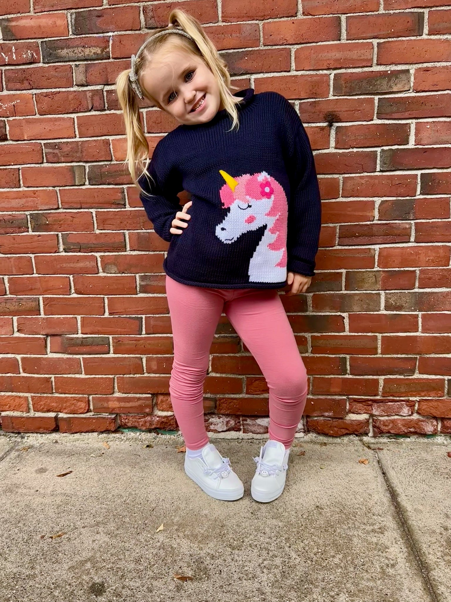 Child wearing a navy sweater with a unicorn design, pink leggings, and white shoes against a brick wall.