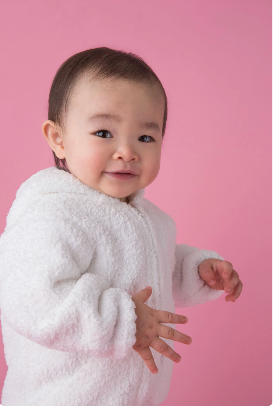 A baby wearing a plush white hooded jacket, standing against a solid pink studio background.