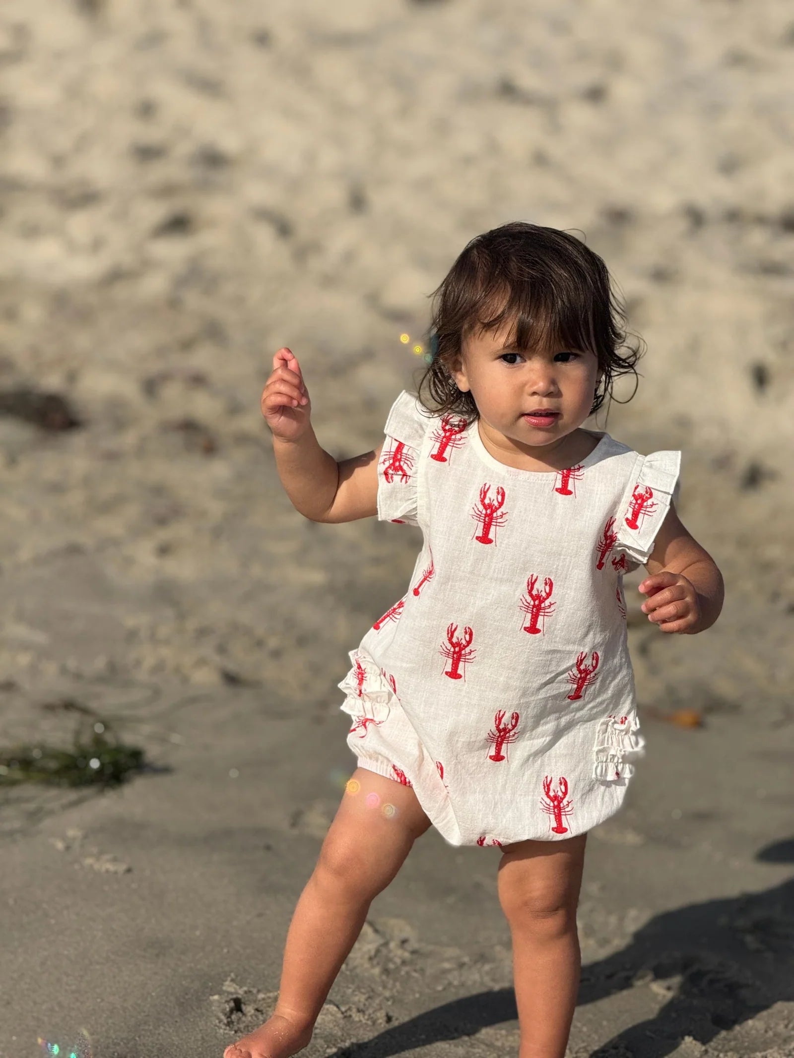 Child in a white dress with red lobster patterns on a sandy beach