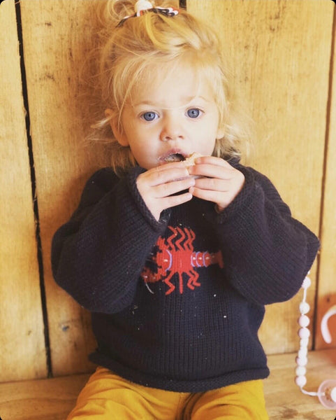 little girl wearing Navy Lobster Roll Neck Sweater eating with crumbs on sweater looking into the camera