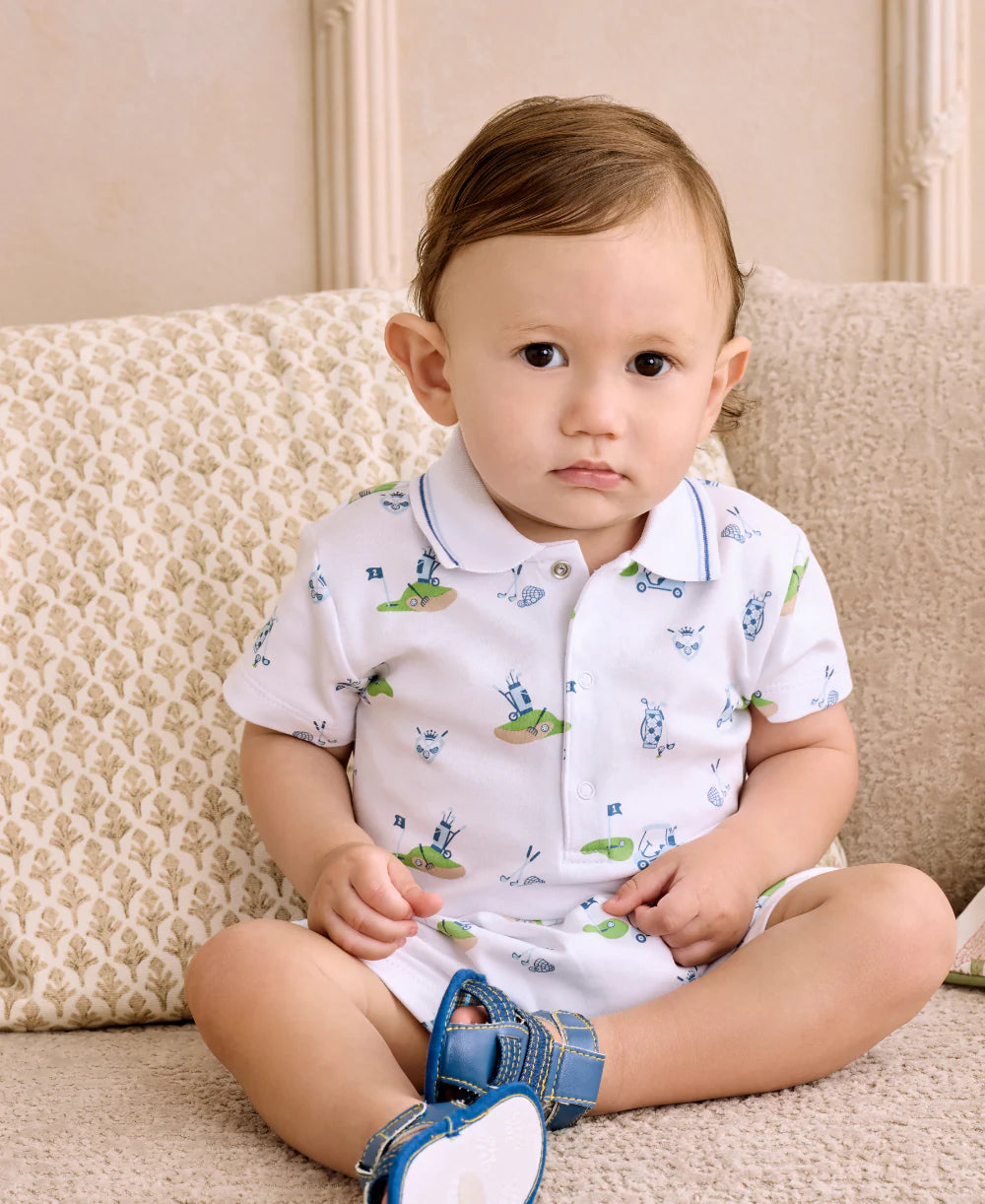 Baby sitting on a couch wearing a white outfit with green and blue patterns.