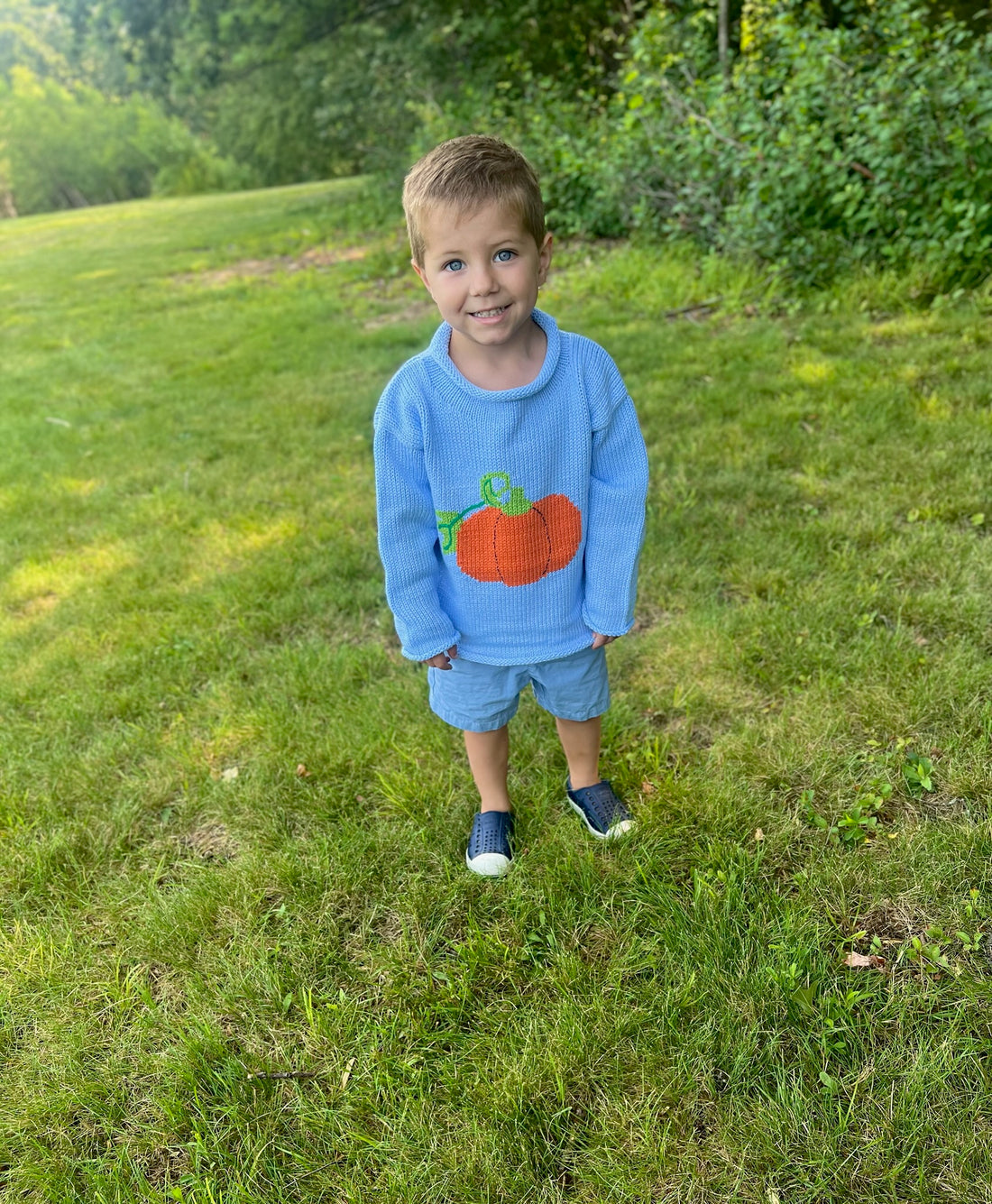 Child wearing a blue sweater with a pumpkin design outdoors on a grassy area