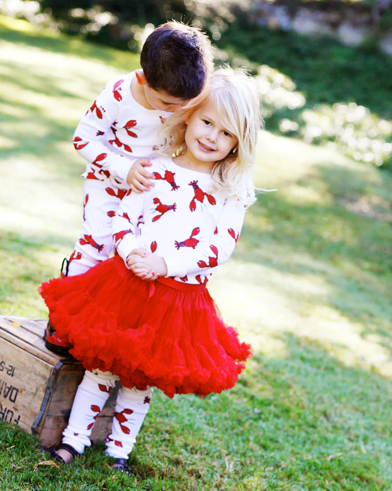 Two young children outdoors on grass wearing matching white outfits with red leaf prints, one with a bright red tutu skirt.
