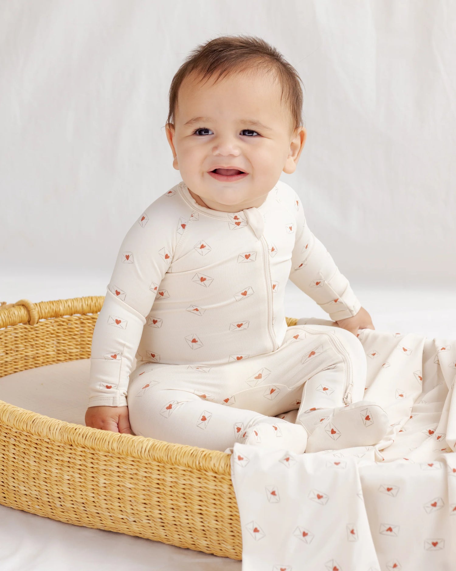 Baby in a white outfit with red patterns sitting in a wicker basket on a light background