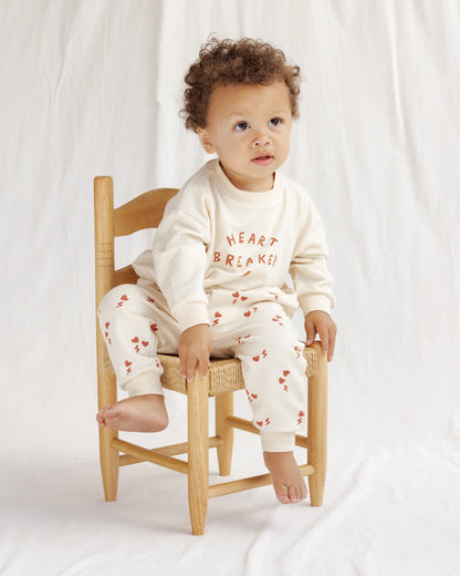 Child wearing a white outfit with text and red patterns, sitting on a wooden chair against a white background