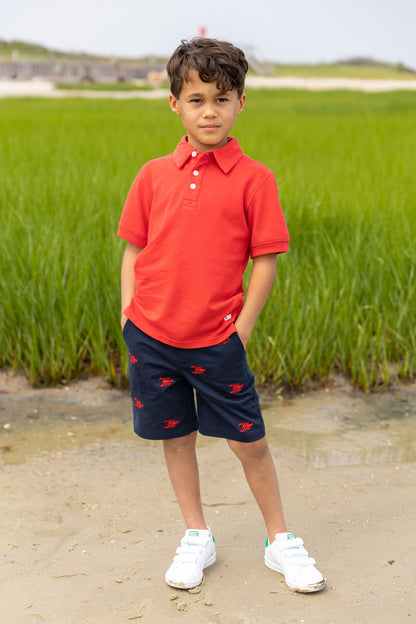 Child wearing a red polo shirt and navy shorts with red accents, standing in a natural setting.