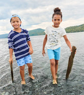 Two children standing on a rocky shoreline with mountains in the background