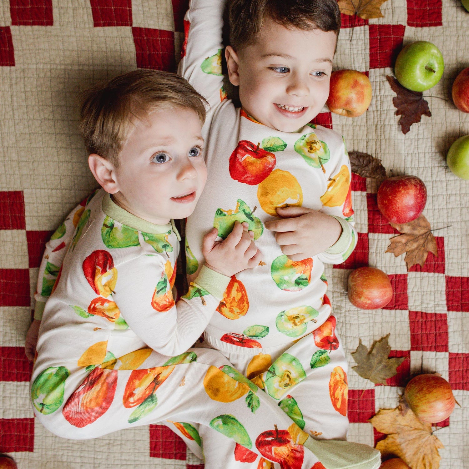 Two children wearing fruit-patterned outfits lying on a checkered blanket with apples and leaves around.