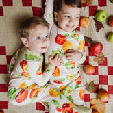 Two children wearing fruit-patterned outfits lying on a checkered blanket with apples and leaves around.