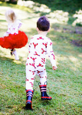 A toddler seen from behind wearing a white one-piece jumpsuit with a red lobster print and red-and-black striped rain boots, walking on a grassy outdoor lawn. Another child in a red tutu is blurred in the background.