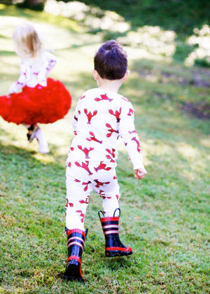 A toddler seen from behind wearing a white one-piece jumpsuit with a red lobster print and red-and-black striped rain boots, walking on a grassy outdoor lawn. Another child in a red tutu is blurred in the background.