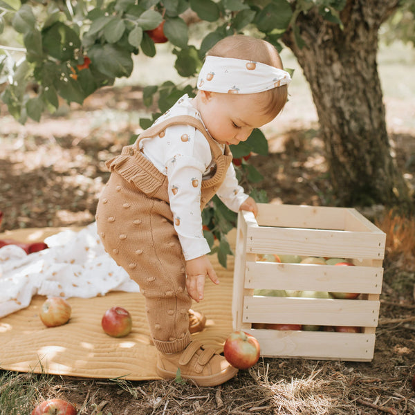 long sleeve apple onesie underneath a sweater knit tan overalls with pom poms