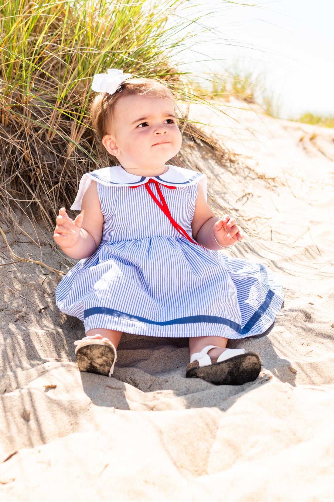 Baby in a blue dress with white shoes sitting on sand dunes.