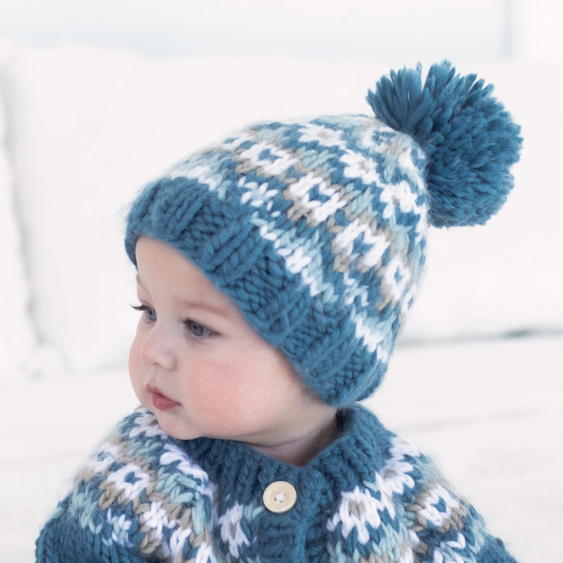 Child wearing a blue knitted hat with a pom-pom and matching cardigan against a light background