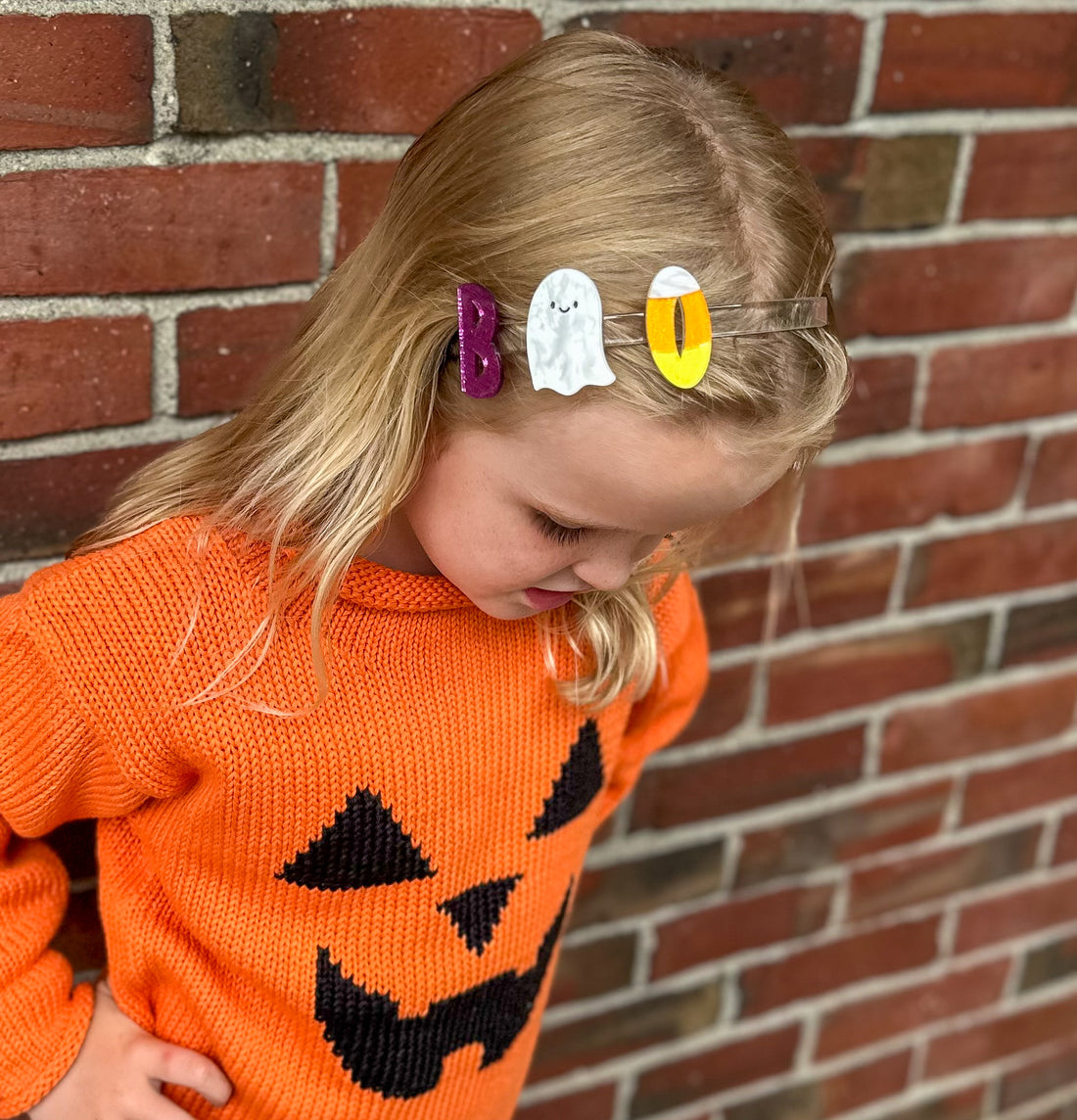 Child wearing an orange sweater with a pumpkin design, standing against a brick wall.