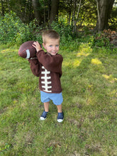 Child wearing a football-themed sweater holding a football outdoors in a grassy area with trees.