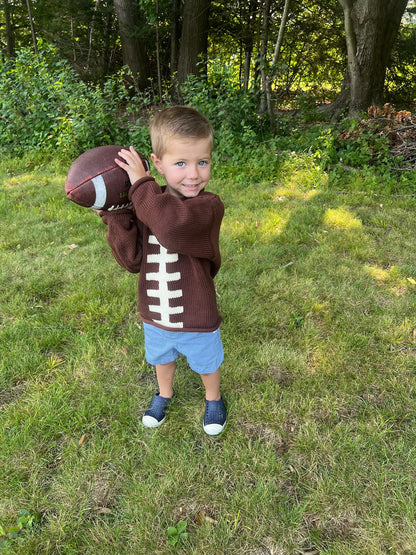 Child wearing a football-themed sweater holding a football outdoors in a grassy area with trees.
