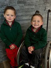 Two young boys, one standing and one in a wheelchair, wearing matching green jackets and red pants against a stone wall.